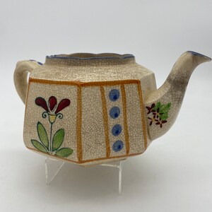 May include: A white ceramic teapot with a crackled glaze and hand-painted floral designs in red, green, and blue. The teapot has a octagonal body and a spout on the right side.