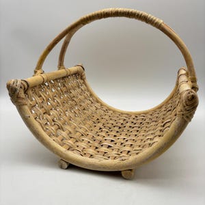 May include: A natural woven basket with a curved design and a large handle. The basket is made of light brown rattan and has a woven pattern. The basket is supported by small wooden feet.