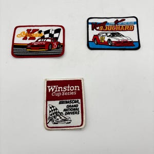 May include: Three rectangular embroidered patches featuring NASCAR themes. One patch shows a red race car with the text Schrader, another displays a white and red race car with Kerry Bouchard 1988 Rookie of the Year, and the third is a red and white Winston Cup Series patch.