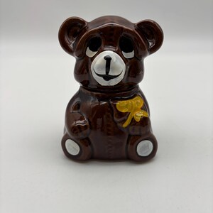 May include: A brown ceramic bear-shaped jar with a yellow bow. The bear has black eyes, white accents, and a white face. The jar is likely for honey or other food items.