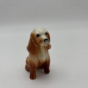 May include: A ceramic figurine of a dog, featuring a seated pose. The dog has a white head with brown ears and markings, a black nose, and brown and tan body. The figurine is detailed and appears to be hand-painted.