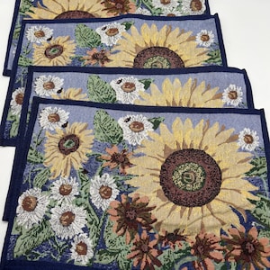 May include: Four woven placemats with a floral design. Each placemat features a large yellow sunflower with a brown center, surrounded by white daisies and other flowers on a blue background. The edges are trimmed with a dark blue border.