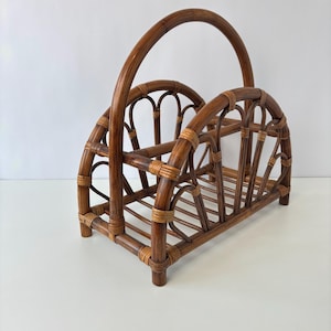 May include: A brown rattan magazine rack with a curved handle. The rack features a rectangular base and arched sides with decorative woven details. The natural wood tones and open design create a vintage aesthetic.