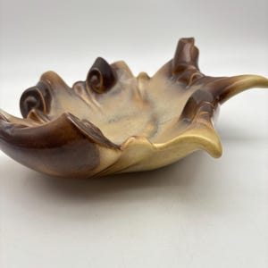 May include: A ceramic leaf-shaped dish with a glossy finish. The dish features a blend of brown and tan hues, with darker brown accents on the edges and curled details. The dish has a shallow bowl shape and a smooth, curved design.