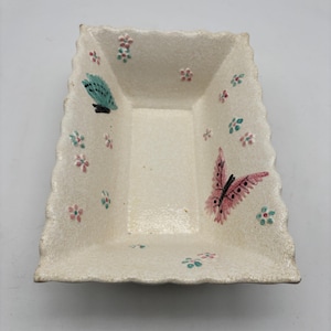 May include: Rectangular off-white ceramic dish with a textured surface and scalloped edges. The dish is decorated with pink and teal butterflies and small pink and teal flowers. The dish is empty.