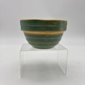 May include: A green and tan ceramic mixing bowl with horizontal ridges. The bowl has a wide, open top and a rounded base. The exterior is a mottled green, while the rim and a band around the middle are tan.