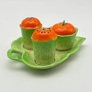 May include: A ceramic salt and pepper set featuring a leaf-shaped tray and three pieces. The salt and pepper shakers are shaped like tomatoes and lettuce. The set is painted in shades of green and orange.