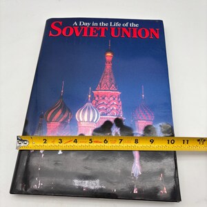 Puede incluir: Un libro titulado "A Day in the Life of the Soviet Union" con una portada que presenta una imagen nocturna de la Catedral de San Basilio. El libro mide aproximadamente 28 cm de largo.