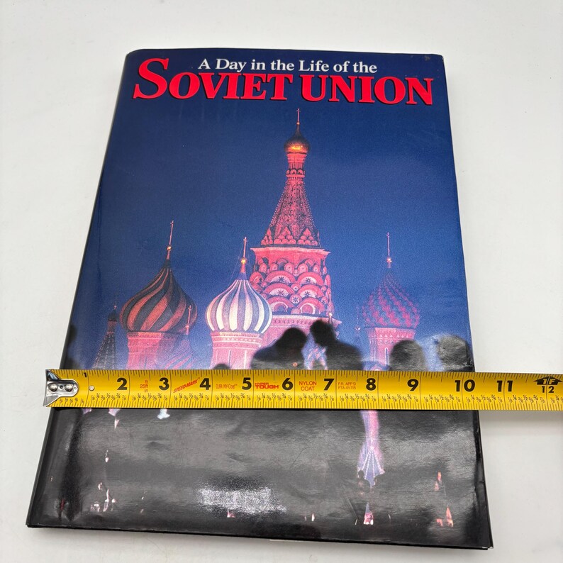 Puede incluir: Un libro titulado "A Day in the Life of the Soviet Union" con una portada que presenta una imagen nocturna de la Catedral de San Basilio. El libro mide aproximadamente 28 cm de largo.
