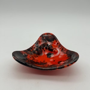 May include: A red and black ceramic ashtray with a unique, abstract design. The ashtray is shaped like a crescent moon and has a glossy finish.