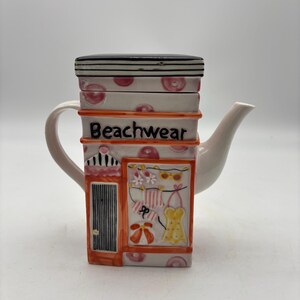 May include: A ceramic teapot shaped like a small shop with a black lid, white walls featuring pink polka dots, and an orange frame. The front displays the text Beachwear above a storefront window showing illustrations of clothing and accessories.
