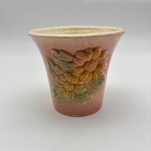 May include: A pink ceramic vase with embossed floral designs. The vase features two large flowers with yellow and orange centers, surrounded by green leaves. The rim is a cream color.