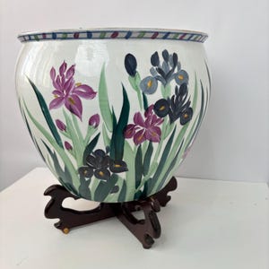 May include: A white ceramic planter with a hand-painted floral design featuring purple, dark purple, and gray irises with green leaves. The planter sits on a dark brown wooden stand. The rim has a striped pattern in blue, green, and purple.