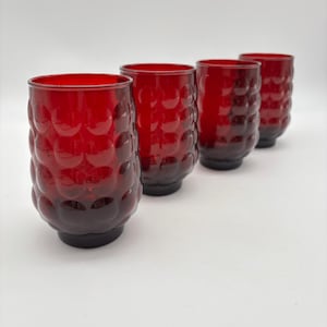 May include: Four red drinking glasses with a textured, bubble-like pattern. The glasses have a dark red base and a lighter red upper portion. The glasses are arranged in a row on a white surface.