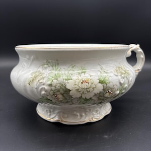 May include: White ceramic bowl with a floral design. The bowl features a detailed embossed floral pattern with white and green flowers. The rim and base are accented with gold trim. The bowl has a handle on one side.