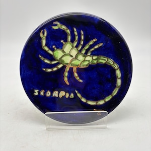 May include: A round, blue ceramic plate featuring a green scorpion illustration with orange accents. The word "SCORPIO" is written in gold lettering. The plate is displayed on a clear stand.