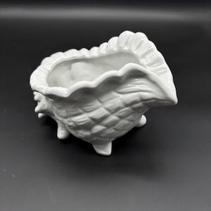 May include: A white ceramic seashell-shaped planter with a textured surface and three small feet. The planter has a scalloped rim and a smooth interior, perfect for small plants or decorative items.