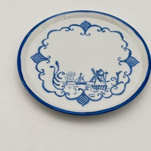 May include: A round, white ceramic plate with a blue rim and a blue decorative border. The center features a blue illustration of a windmill, boat, and landscape scene. The plate is approximately 6 inches in diameter.