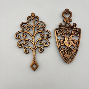 May include: Two ornate, copper-colored metal trivets. One is tree-shaped with intricate scrollwork, and the other is shield-shaped with a heart and bird design. Both have a vintage aesthetic.