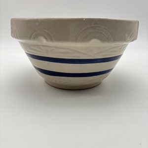 May include: A beige ceramic mixing bowl with two navy blue stripes. The bowl has a wide rim with a decorative embossed pattern. The bowl is likely vintage or antique.