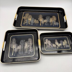 May include: A set of three black rectangular serving trays with rounded corners and gold-toned rims. Each tray features a central graphic of a forest with gold and white trees against a dark blue striped background and light-colored woven handles.