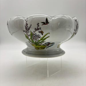 May include: A white porcelain bowl with a scalloped rim and a pedestal base. The exterior features a colorful botanical illustration of green plants, purple flowers, a small brown butterfly, and a dark bird in flight against a white background.