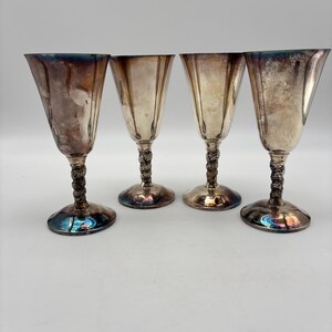 May include: Four vintage silver goblets with a fluted design. The goblets have a decorative stem and a wide base. The silver has a tarnished appearance, with hints of blue and brown. The goblets are likely used for serving wine or other beverages.