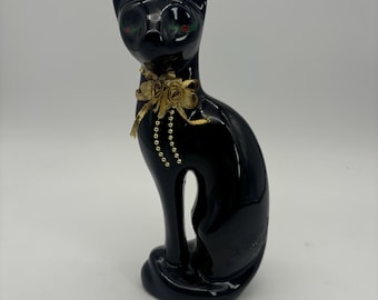May include: Black ceramic cat figurine with gold accents. The cat is sitting with its tail curled around its legs. It has a gold bow with a small flower design around its neck.