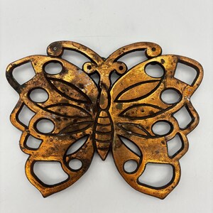 May include: A butterfly-shaped trivet crafted from metal with a distressed, aged gold-tone finish. The design features an openwork pattern with cutouts throughout the wings and a segmented body, showcasing a weathered texture across the surface.