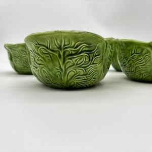 May include: A set of three green ceramic bowls, each featuring a molded, raised texture that mimics the appearance of overlapping cabbage leaves. The bowls have a glossy finish and a scalloped, uneven rim, with one bowl centered in the foreground.