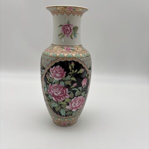 May include: A tall, decorative ceramic vase with a cream-colored neck and a flared rim. The vase features a black central panel with pink roses and green leaves. The design includes a patterned border and a cream-colored base.