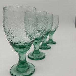May include: A set of four light green glass goblets arranged in a row. Each goblet features a textured, embossed surface pattern resembling clusters of grapes on the bowl, a short stem, and a circular base with a matching textured edge.