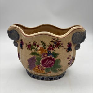 May include: A ceramic planter with a cream-colored crackle glaze finish, featuring a colorful floral pattern of roses and grapes. The vessel has a wavy rim, two blue scroll-shaped side handles, and a blue floral border around the base.