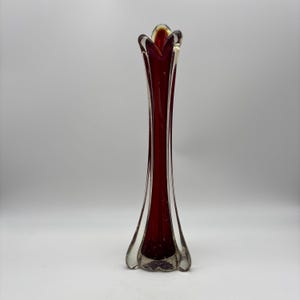 May include: A tall, slender, ruby-red glass vase with a flared base and a flower-like top. The vase has a clear glass exterior, with the red interior visible through the clear glass. The vase is approximately 12 inches tall.