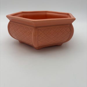May include: A terracotta-colored ceramic planter with a geometric, hexagonal shape. The planter features a diamond-patterned texture on its curved sides and a slightly flared rim. The base has small feet, and the interior is smooth.