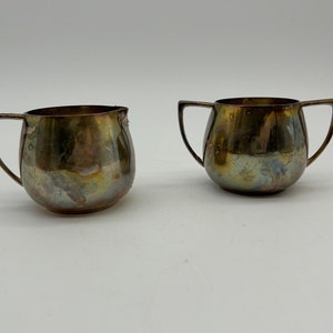 May include: Two silver creamer or sugar bowls with a rounded body and two handles. The bowls have a worn, aged look.