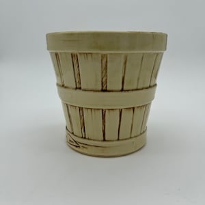 May include: A cream-colored ceramic planter shaped like a woven basket with brown accents. The planter has a wide opening and a rounded bottom.