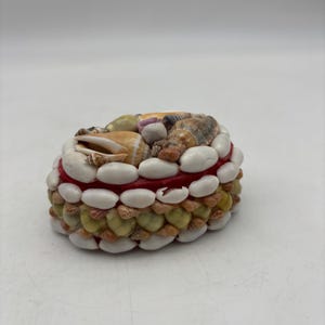 May include: A round, decorative box covered in various seashells. The box features a red band in the middle, with white and tan shells arranged around the sides. The top is adorned with a variety of shells in different colors and sizes.