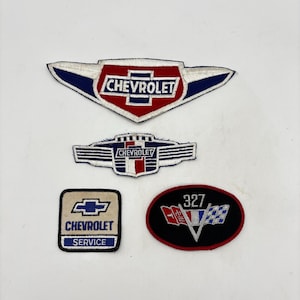 May include: Four embroidered fabric patches featuring Chevrolet branding. The top patch is wing-shaped with red, white, and blue. The middle patch is a blue and white shield. The bottom left is a square Chevrolet Service patch. The bottom right is an oval 327 patch.