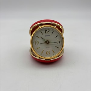 May include: A vintage red and gold travel alarm clock. The clock face is white with black numerals and hands, and the brand name "Linden" is printed on the face. The clock is encased in a red, rounded case that opens to reveal the clock.