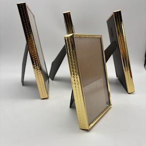 May include: Four rectangular picture frames with gold-colored borders and black easel backs. The frames are arranged at different angles, showcasing their design. The gold borders have a textured pattern, adding detail to the frames.
