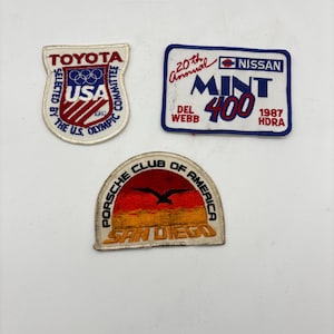 May include: Three embroidered patches: a shield-shaped Toyota 1980 USA Olympic Committee patch, a rectangular 20th Annual Nissan Mint 400 Del Webb 1987 HDRA patch with blue border, and a semi-circular Porsche Club of America San Diego patch.