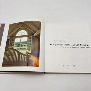 May include: An open book with a photograph of a bedroom with a balcony and a bed. The text on the right page reads "Joan Kohn's it's your bed and bath Hundreds of Beautiful Design Ideas". The publisher is Bulfinch Press.