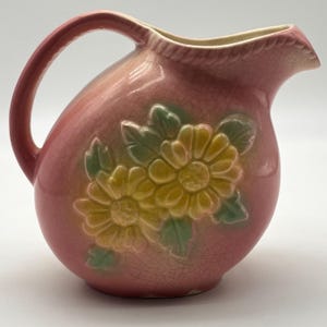 May include: Pink ceramic pitcher with a handle and spout. The pitcher features embossed yellow flowers with green leaves. The pitcher has a crackle glaze finish and a light pink to peach color gradient.