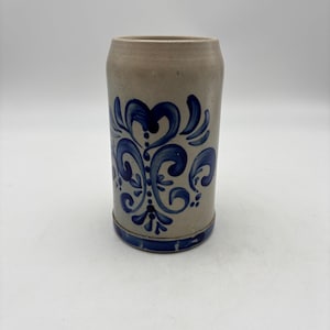 May include: A cylindrical stoneware beer mug featuring a light gray body with a hand-painted cobalt blue floral pattern. The design includes symmetrical scrollwork and flourishes, with a solid blue band encircling the base of the vessel.