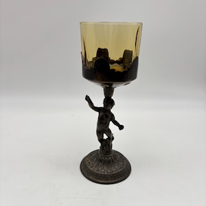 May include: An antique candle holder with a bronze cherub base supporting a yellow-tinted glass cup. The cup has a decorative rim. The base is round with an ornate design.