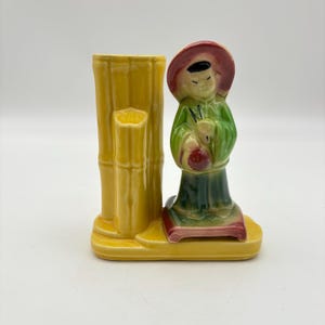 May include: A vintage ceramic vase featuring a yellow bamboo-style column and a figurine of a person in traditional attire. The figurine wears a red hat and green clothing, holding a red object. The base is yellow with red accents.