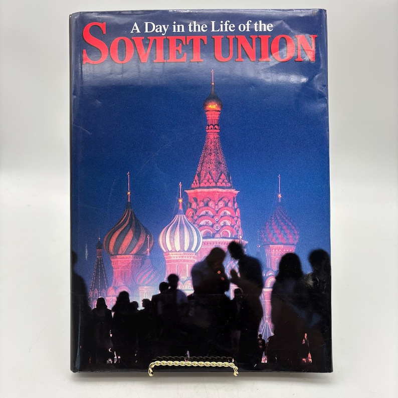 Puede incluir: Un libro de tapa dura titulado "A Day in the Life of the Soviet Union". La portada presenta una imagen nocturna de la Catedral de San Basilio en Mosc&uacute;, con siluetas y texto rojo.