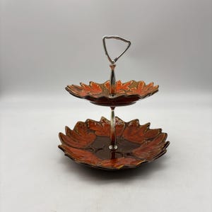 May include: Two-tiered serving tray with a silver handle and stem. The trays are shaped like leaves and are glazed in shades of orange and brown. The tray is designed for serving desserts or appetizers.
