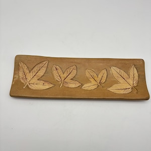 May include: A rectangular, tan-colored ceramic tray with four embossed leaf designs. The leaves are detailed with veining and have a lighter brown color than the tray. The tray has a slightly curved edge and is set against a white background.